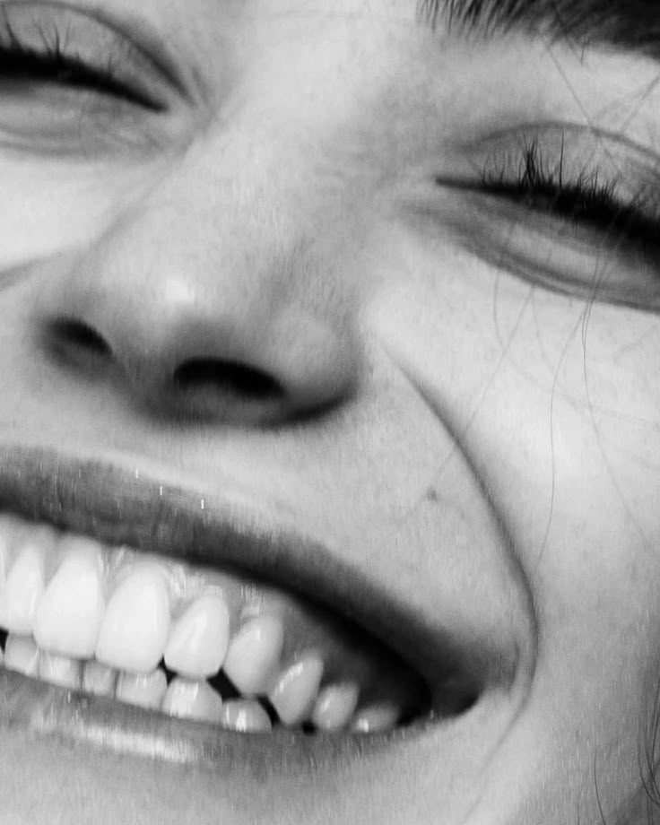 Pessoa idosa segurando um cartão com um grande sorriso impresso em frente à boca, criando um efeito visual divertido; usa óculos e veste roupa clara com bordado floral, em frente a uma cortina simples.